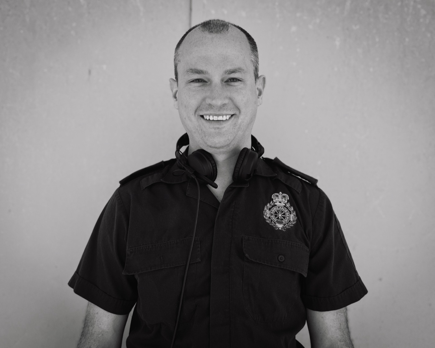 Black and white portrait of Michael smiling, in an NHS Ambulance Service shirt with headphones round his neck.