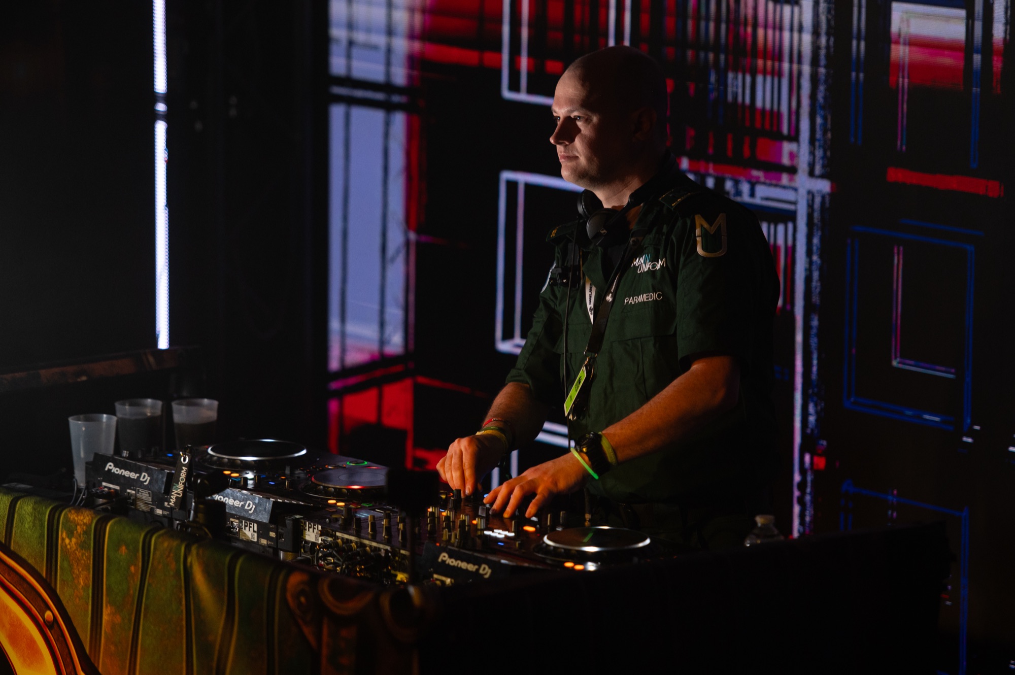 Michael in paramedic uniform with his hands on a Pioneer DJ mixer, in front of a red and blue video wall.